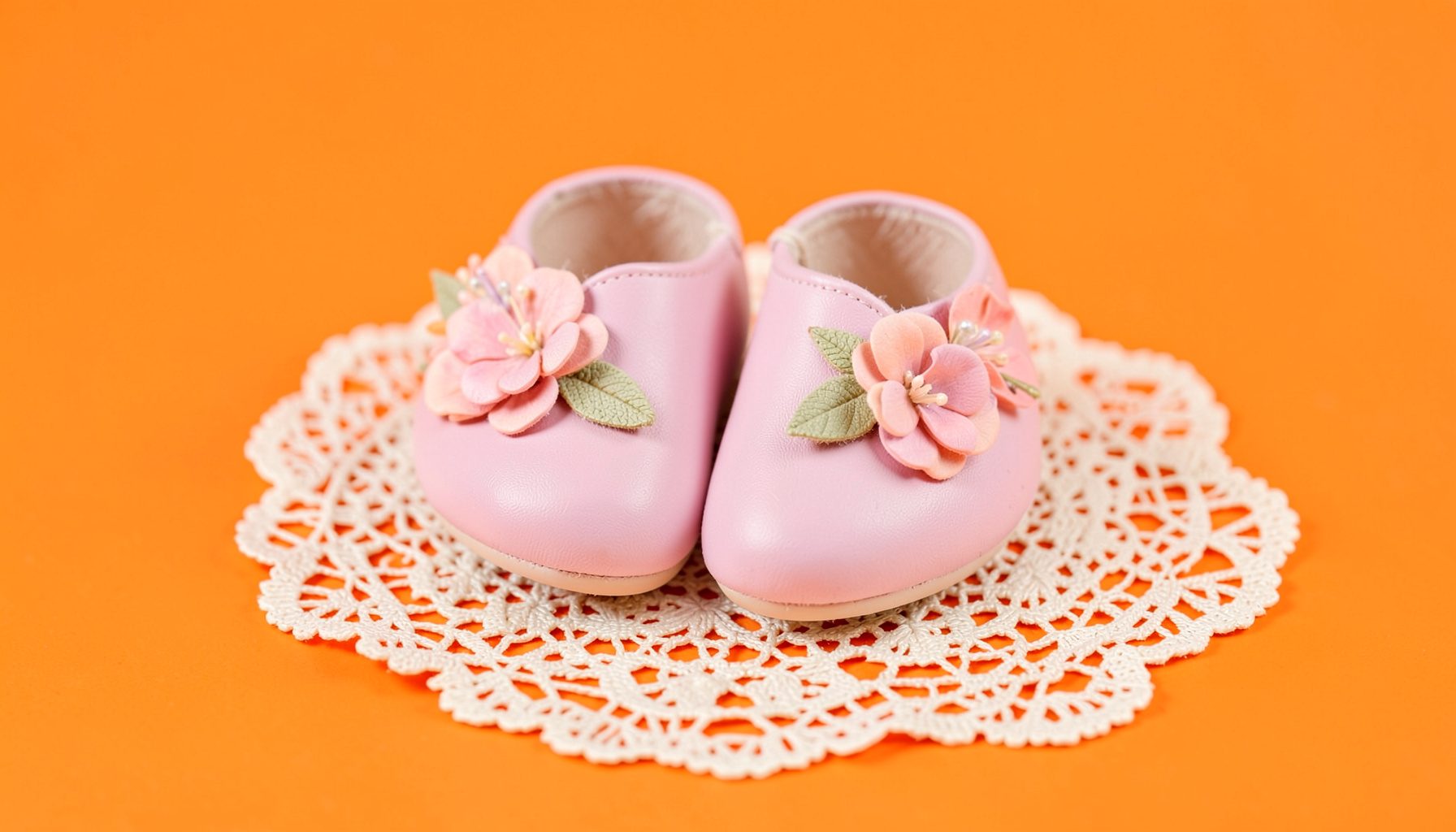 Delicate pastel-hued leather baby shoes with floral embellishments arranged on a vintage lace doily, soft pink and cream tones dominating the scene.