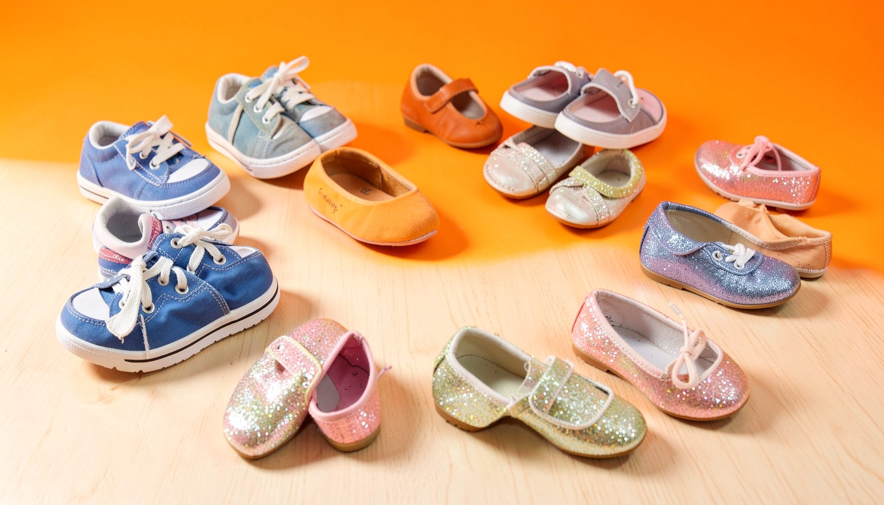 A bright, airy flat lay of colorful children's shoes — tiny canvas sneakers, soft leather sandals, and glittery ballet flats — scattered playfully across a pale wooden floor, with warm natural light casting gentle shadows.