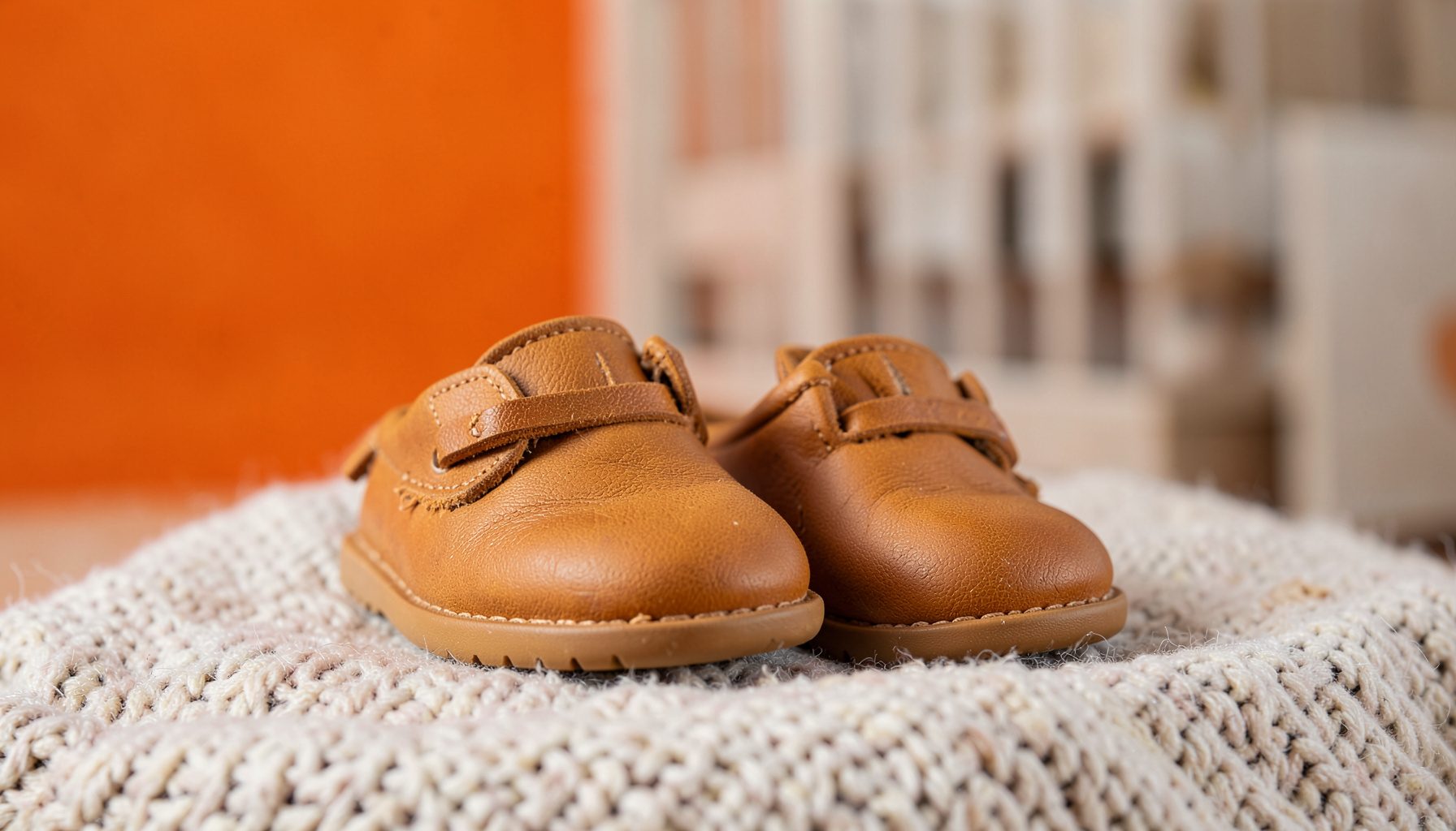 A pair of soft leather toddler shoes in warm caramel brown resting on a textured wool blanket, with a muted Scandinavian-style nursery blurred in the background.