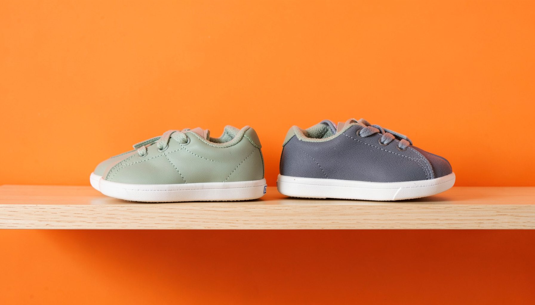 Minimalist children's sneakers in muted sage green and slate gray lined up on a light oak shelf, with clean Nordic design elements and soft diffused daylight.