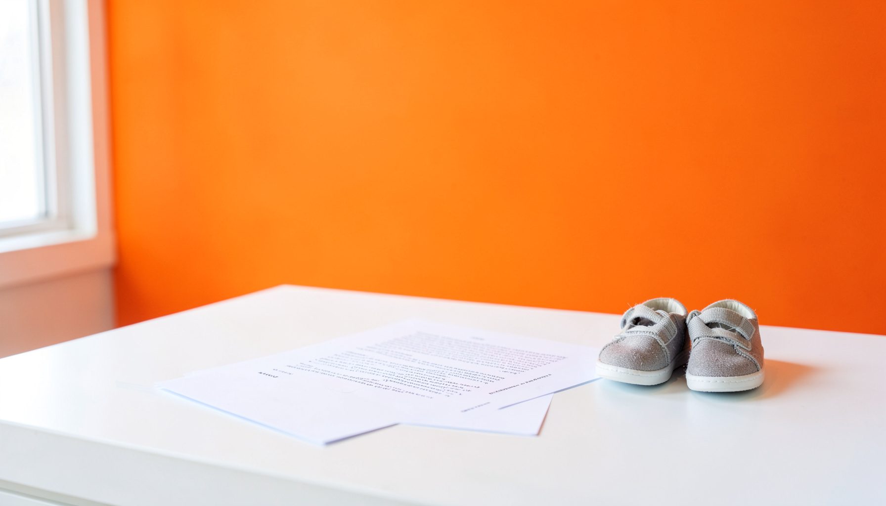 A clean white desk with a printed document and a pen beside a small pair of children's shoes, soft natural light from a window, calm and orderly mood.